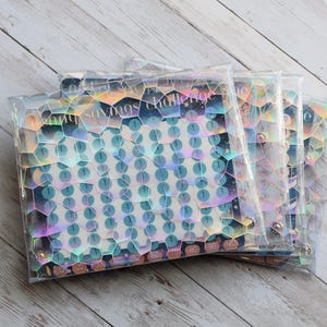 May include: Several clear, iridescent savings challenge pouches are stacked on a light wood surface. Each pouch has a honeycomb pattern and contains a savings tracker with teal circles and the text "savings challenge".
