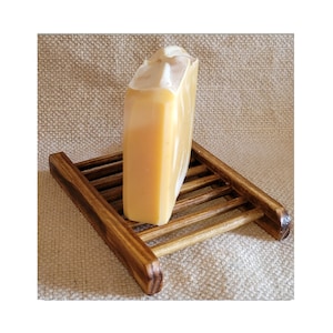 May include: A rectangular bar of soap with a light yellow hue and white marbling, resting on a small, stained wooden soap dish. The soap dish has a slatted design and is made of dark brown wood. The background is a textured, neutral-colored fabric.