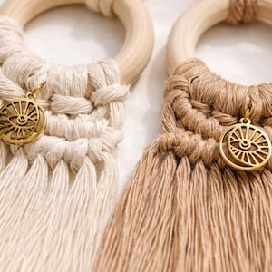 May include: Two macrame wall hangings featuring light wood rings and cotton cord in cream and tan. Each piece includes a gold-toned metal charm shaped like an eye with radiating lines, attached to the knotted cord section above a long fringe.