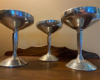 May include: Three silver champagne coupe glasses with a classic design. Each glass features a wide, shallow bowl atop a slender stem and a circular base. The glasses are arranged on a wooden surface, reflecting the light and creating a vintage aesthetic.