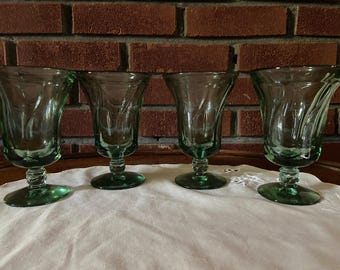 May include: Four green-tinted glass goblets with a tulip-shaped bowl and a decorative stem. The goblets are arranged on a white cloth, with a brick wall in the background. The glasses are approximately 6 inches tall.