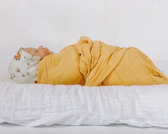May include: A baby swaddled in a mustard yellow muslin wrap, lying on a white pillow. The baby is wearing a white and brown patterned hat with a small fox design.