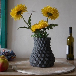 Könnte beinhalten: Eine graue Keramikvase mit geometrischem Muster hält einen Strauß gelber Blumen. Die Vase steht auf einem Holztisch.