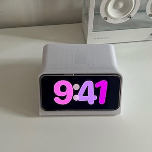 Può includere: Un supporto per telefono bianco con un telefono bianco che mostra l'ora 9:41 in rosa e viola.