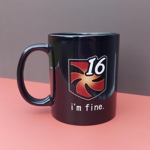 以下が含まれることがあります： 赤い渦巻きと数字の16が描かれた、白と赤のロゴがついた黒いセラミック製のコーヒーカップ。ロゴの下に「i'm fine.」という文字が印刷されています。
