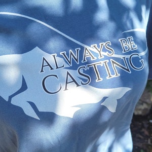 May include: A blue t-shirt with a white fish silhouette and the text "ALWAYS BE CASTING" printed on the front.