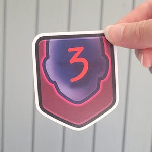 May include: A sticker with a shield shape, featuring a purple and red design with the number 3 in orange. The sticker has a white border and is held by a hand.