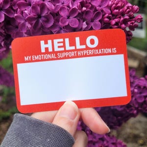 May include: Red name tag with white text that reads "HELLO MY EMOTIONAL SUPPORT HYPERFIXATION IS".