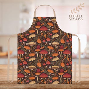 May include: An apron with a dark background and a repeating pattern of mushrooms in shades of orange, red, and beige. The apron has a beige neck strap and ties. The text "Blissful Seasons" is visible on the right side.