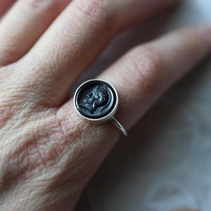 May include: A silver ring featuring a black cameo with a classical profile. The ring's design is a circular, raised relief set in a simple silver band. The ring is shown on a person's finger.