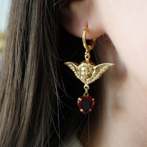 May include: Gold-toned hoop earrings featuring a cherub design with outstretched wings and a red heart-shaped gemstone pendant. The earrings are worn on a person's ear, showcasing the intricate details of the jewelry.