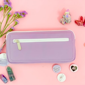 Può includere: Una custodia lilla per Nintendo Switch con cerniera bianca e cinturino da polso. La custodia è su uno sfondo rosa con fiori viola, cristalli e spille decorative. Le spille hanno testo e gesti delle mani.