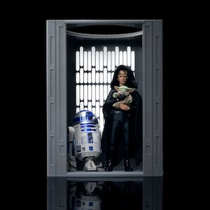 Peut inclure: Un diorama Star Wars avec des figurines de Luke Skywalker tenant Grogu et R2-D2. Les figurines sont dans un décor gris avec un motif en forme de fenêtre en arrière-plan. Luke porte une cape noire et Grogu est dans ses bras.