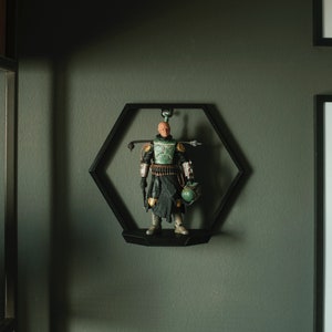 Pode incluir: Uma vitrine hexagonal preta com uma figura de ação Star Wars dentro. A figura está vestindo uma armadura verde e marrom com um capacete e um rifle blaster. A figura está em pé em uma plataforma preta.