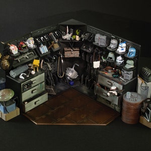 May include: A detailed model of a Star Wars themed workshop featuring various tools, weapons, and helmets. The workshop is constructed from cardboard and painted in a realistic style. The model includes a range of Star Wars characters, including a Mandalorian, a Stormtrooper, and a Jedi.