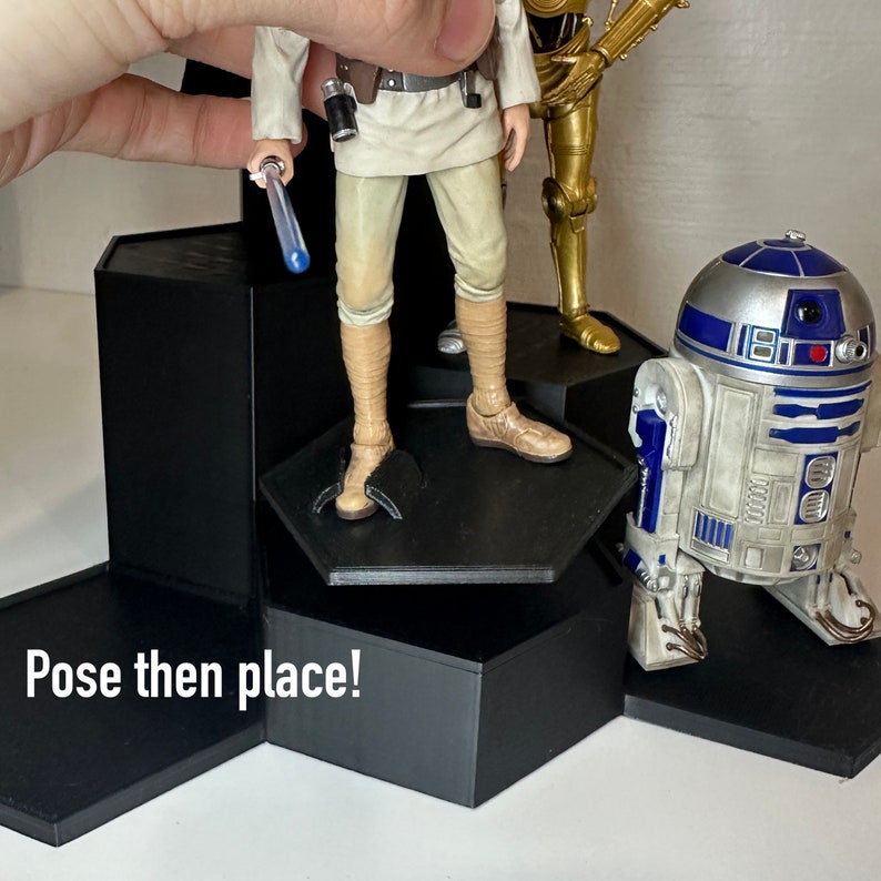 May include: A black hexagonal display stand with three levels, featuring a Star Wars action figure of Luke Skywalker holding a blue lightsaber, a gold C-3PO action figure, and a white and blue R2-D2 action figure. The stand has the text "Pose then place!" printed on the bottom level.