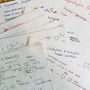 Op de afbeelding: Een verzameling handgeschreven notities op ruitjespapier, met details over organische chemieconcepten. De notities, geschreven met zwarte en rode inkt, behandelen onderwerpen als organometaalverbindingen en aromaticiteit. Het woord "Spectrometer" is zichtbaar.