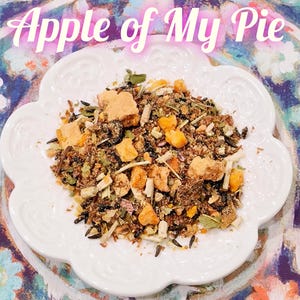 May include: A white, scalloped-edge plate holds a mix of dried ingredients, including small, tan-colored cubes, dark grains, and green herbs. The text "Apple of My Pie" is displayed in a light pink, cursive font above the plate.