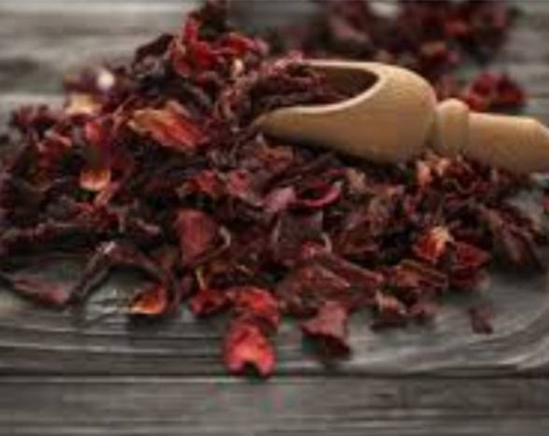 Puede incluir: Primer plano de flores de hibisco secas con una cuchara de madera. Las flores son de color rojo intenso y parecen sueltas y texturizadas. La cuchara es de color marr&oacute;n claro y est&aacute; parcialmente llena de flores. El fondo es una superficie de madera.