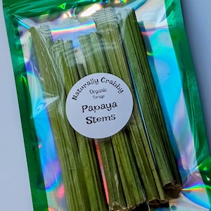 May include: A clear, green, iridescent package containing several green papaya stems. A white circular label reads "Naturally Crabby Organic Forage Papaya Stems". The stems are a vibrant green color and appear to be dried.