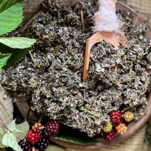 May include: A close-up of a bowl filled with dried green herbs, with a wooden spoon scooping out the herbs. The bowl is on a brown surface, and there are some blackberries scattered around the bowl.