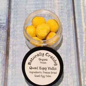 May include: A small clear jar filled with freeze-dried quail egg yolks. The jar has a black lid with the text "Naturally Crabby Organic Protein Quail Egg Yolks Ingredients: Freeze Dried Quail Egg Yolks".