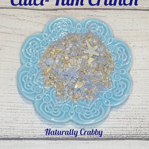 May include: A blue and white ceramic dish with a scalloped edge, filled with a mixture of dried herbs, sea shells, and starfish. The text "Calci-Yum Crunch" is at the top of the image and "Naturally Crabby" is at the bottom.