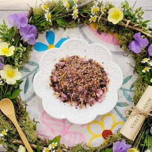 May include: A white, scalloped-edge plate holds a mix of dried herbs and flowers, surrounded by a floral wreath. The wreath includes purple sweet peas, yellow buttercups, and white daisies. The text "Violet Meadow Mix" is visible.