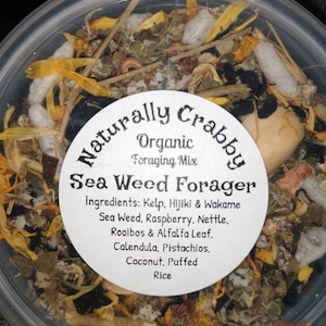 May include: A clear plastic container filled with a variety of dried ingredients, including seaweed, kelp, and other herbs. The label reads "Naturally Crabby Organic Foraging Mix Sea Weed Forager" and lists the ingredients: Kelp, Hijiki & Wakame Sea Weed, Raspberry, Nettle, Rooibos & Alfalfa Leaf, Calendula, Pistachios, Coconut, Puffed Rice.