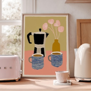 Może przedstawiać: Różowy toster z chromowanymi detalami stoi obok oprawionego wydruku przedstawiającego kawiarkę, kubki i kwiaty. Na drewnianym blacie widoczny jest również kremowy czajnik elektryczny i mały biały kubek.