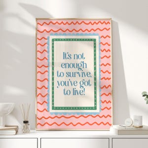 Pode incluir: Uma impressão artística emoldurada com fundo rosa e padrão ondulado. A impressão apresenta o texto "It's not enough to survive, you've got to live!" em azul. A moldura é de cor clara.