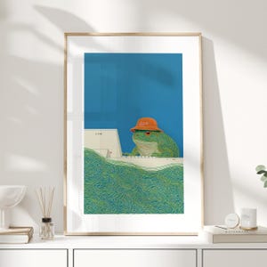 Op de afbeelding: Een ingelijste kunstdruk met een groene kikker met een oranje hoed, die op een computer werkt. De achtergrond is blauw en de voorgrond heeft groene, wervelende lijnen. Op de hoed staat het woord "GOOD".