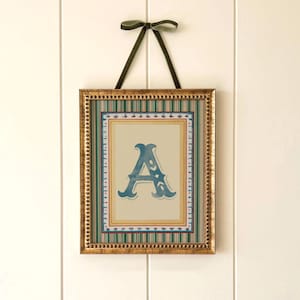 Può includere: Stampa incorniciata con la lettera "A" in blu. La cornice dorata circonda un bordo a righe verdi e bianche. Un nastro verde è legato in alto per appenderla. Decorazione murale.