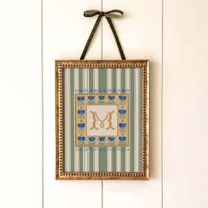 Puede incluir: Un bordado en punto de cruz enmarcado con la letra "M" en un marco dorado. La obra de arte tiene un fondo a rayas verdes y blancas, un borde floral y una letra dorada. Se utiliza una cinta verde oscuro para colgar, mide unos 30 x 20 cm.