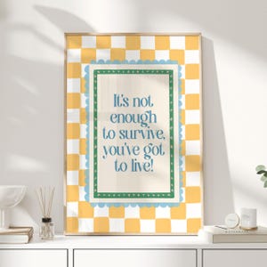 Pode incluir: Uma impressão emoldurada com o texto "It's not enough to survive, you've got to live!" em azul. A impressão tem uma borda xadrez amarela e branca, uma borda ondulada azul e uma borda interna verde. A moldura é de cor de madeira clara.