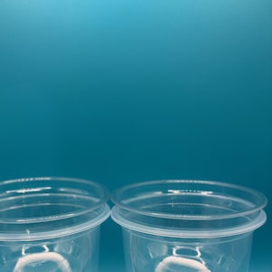 May include: Two clear plastic cups with white wicks inside. The cups are sitting on a blue surface.