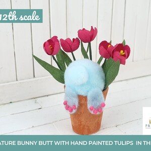 Op de afbeelding: Een miniatuur konijnenkontje figuur dat in een terracotta pot zit met handgeschilderde roze tulpen. Het konijn is lichtblauw met roze poot afdrukken. De pot is 1:12 schaal.