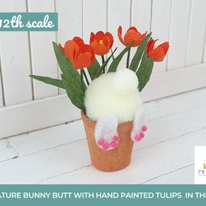 Könnte beinhalten: Ein Miniatur-Hasenhintern in Weiß mit rosa Füßen sitzt in einem Terrakotta-Topf mit handbemalten orangen Tulpen. Der Topf steht auf einer weißen Holzoberfläche. Maßstab 1:12.
