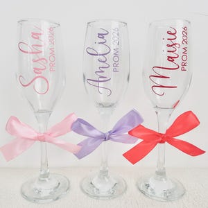 May include: Three clear glass champagne flutes, each personalised with a name in cursive script and the text PROM 2026. The first glass features the name Sasha in pink with a light pink ribbon, the second Amelia in purple with a lavender ribbon, and the third Maisie in red with a red ribbon tied around the stem.