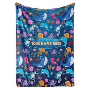 May include: A blue fleece blanket with a colorful ocean-themed pattern. The pattern includes whales, dolphins, sharks, jellyfish, seahorses, starfish, and seashells. The text "YOUR NAME HERE" is printed in a white oval on the blanket.