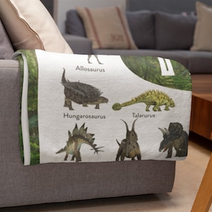 May include: A white throw blanket with a green leafy background and illustrations of different dinosaurs. The dinosaurs are labeled with their names: Allosaurus, Hungarosaurus, Talarurus, and two others.