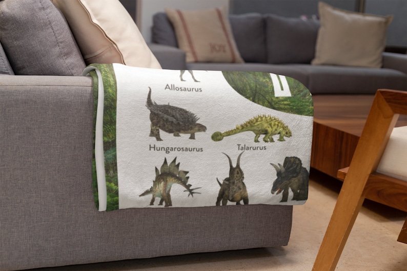 May include: A white throw blanket with a green leafy background and illustrations of different dinosaurs. The dinosaurs are labeled with their names: Allosaurus, Hungarosaurus, Talarurus, and two others.