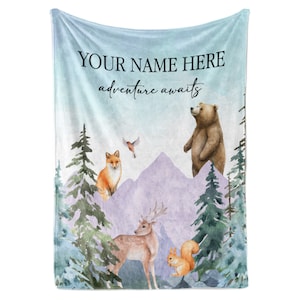 May include: A blue fleece blanket with a watercolor design of a bear, fox, deer, and squirrel in a forest setting. The text "YOUR NAME HERE adventure awaits" is printed on the blanket.