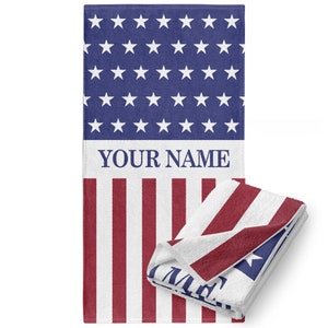 May include: A beach towel with a red, white, and blue American flag design. The top half of the towel features a blue field with white stars. The bottom half of the towel features alternating red and white stripes. The text "YOUR NAME" is printed in blue on the towel.