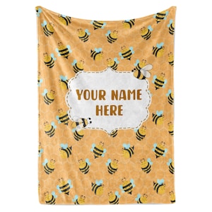 May include: A yellow fleece blanket with a pattern of bees and honeycombs. The blanket has a white label with the text "YOUR NAME HERE".