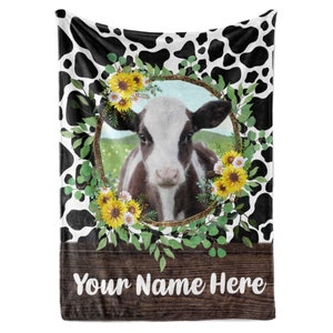 May include: A black and white cow with a brown spot on its face is surrounded by a wreath of sunflowers and greenery. The wreath is on a black and white cow print background. The text "Your Name Here" is on a brown wooden sign at the bottom of the image.
