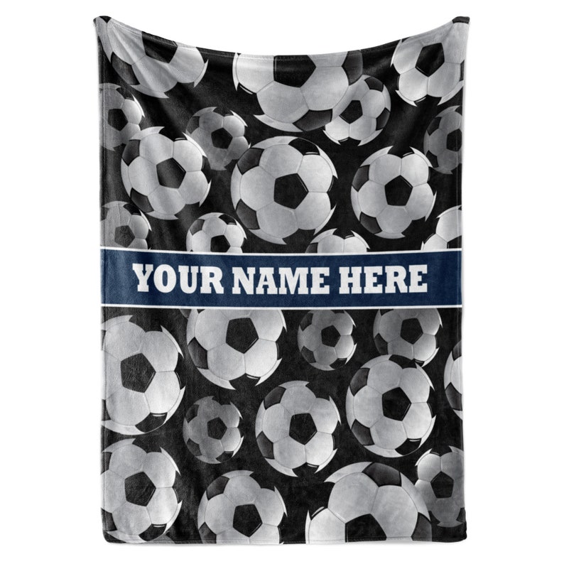 May include: A black and white soccer ball patterned blanket. The blanket features a repeating pattern of soccer balls in various sizes. A blue banner across the blanket reads "YOUR NAME HERE" in white text.