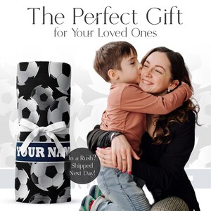 May include: A rolled black blanket with a white soccer ball pattern, tied with a white ribbon and a blue band that says "YOUR NAME" in white. The text "The Perfect Gift for Your Loved Ones" is at the top.
