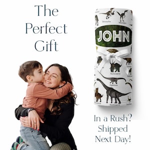 May include: A white blanket with a dinosaur print and the name "JOHN" in green text. The blanket is rolled up and tied with a white ribbon. A mother and son are hugging in the background.