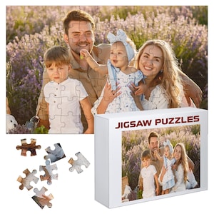 May include: A family portrait jigsaw puzzle with a white box. The puzzle features a family of four in a lavender field. The box has the words "JIGSAW PUZZLES" in red. Several puzzle pieces are scattered around the box.